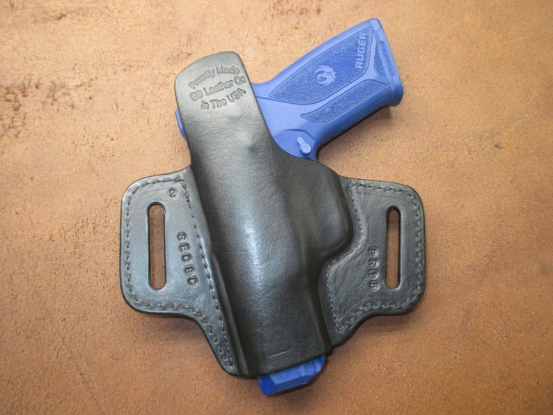 Może przedstawiać: Czarna sk&oacute;rzana kabura z niebieskim pistoletem Ruger w środku. Kabura ma pętlę na pasek i jest oznaczona napisem "Proudly Made by GG Leather Co. In The USA".