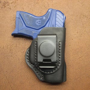 May include: A black leather holster with a metal clip holds a blue handgun. The holster is stitched with black thread and has a circular clip. The handgun is a compact size.