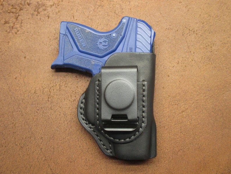 May include: A black leather holster with a metal clip holds a blue handgun. The holster is stitched with black thread and has a circular clip. The handgun is a compact size.