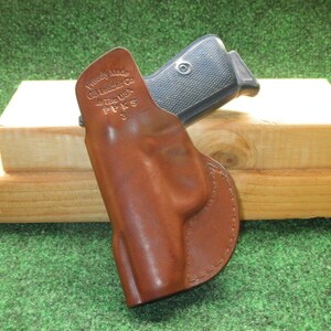 May include: A brown leather holster with a firearm inside. The holster is stamped with the text "Proudly Made GB Leather Co in The USA PPKS 3". The firearm has a black grip. The background is a wooden block and green surface.