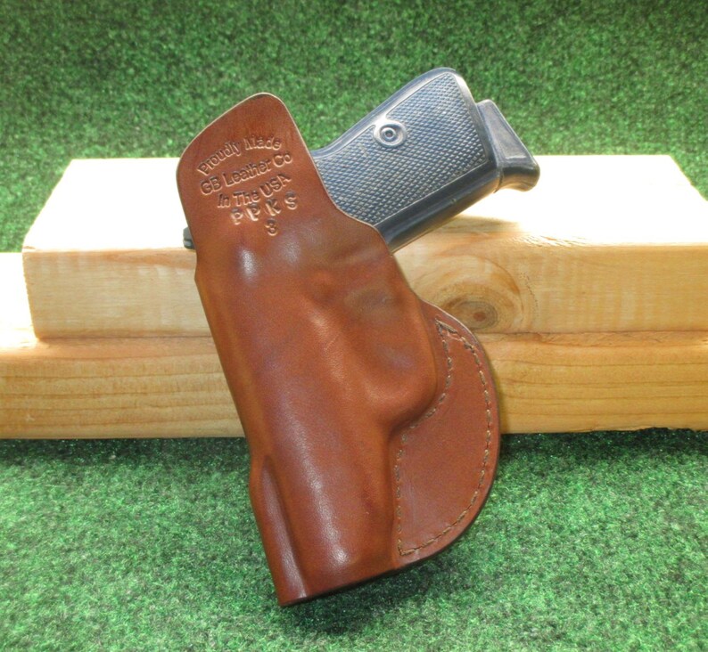 May include: A brown leather holster with a firearm inside. The holster is stamped with the text "Proudly Made GB Leather Co in The USA PPKS 3". The firearm has a black grip. The background is a wooden block and green surface.