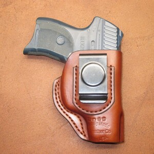 May include: A brown leather holster with a silver metal clip, holding a black handgun. The holster is stitched with a lighter brown thread and has the text "Eoos Family Made Leather Co USA" embossed on the front.