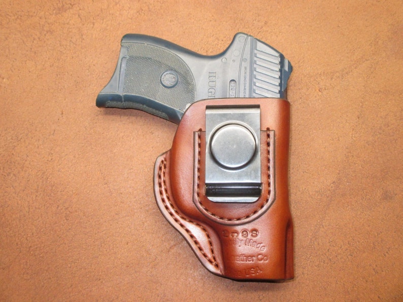May include: A brown leather holster with a silver metal clip, holding a black handgun. The holster is stitched with a lighter brown thread and has the text "Eoos Family Made Leather Co USA" embossed on the front.