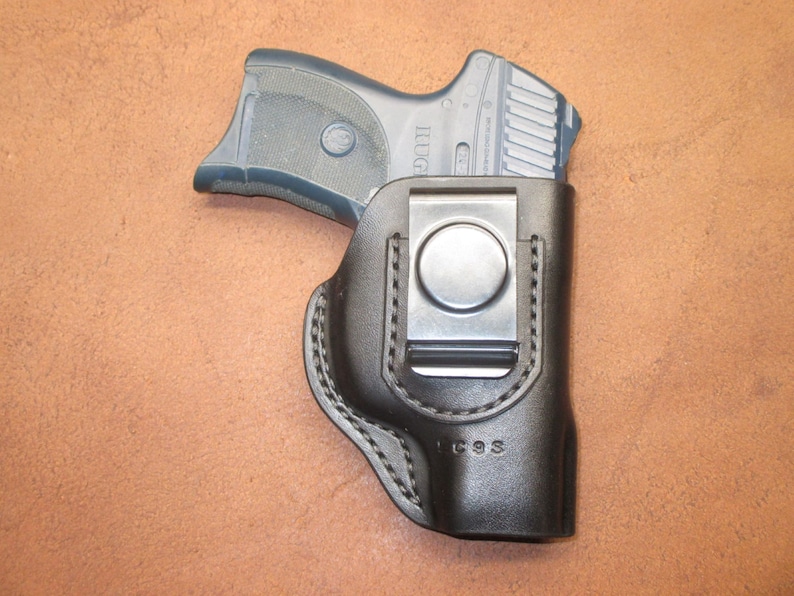 May include: A black leather holster with a silver metal clip, holding a black and gray handgun. The holster is stitched with white thread and has the letters "LC9S" stamped on it. The handgun is partially visible.
