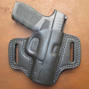 May include: A black leather holster with a black handgun inside. The holster has a molded design with visible stitching around the edges and belt loops. The background is a brown, textured surface.