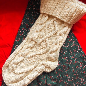 May include: A cream-colored knitted stocking with a cable knit pattern. The stocking is laying on a red and green fabric with a candy cane pattern.