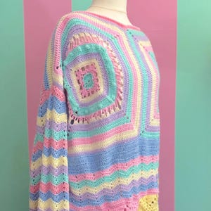Puede incluir: Un suéter de ganchillo de colores pastel exhibido en un maniquí. El suéter presenta un diseño de patchwork con cuadrados y rayas en tonos de rosa, azul, amarillo y lavanda. Las mangas son largas y el estilo es holgado.