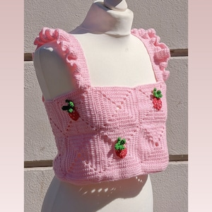 Könnte beinhalten: Ein rosa gehäkeltes Crop-Top mit quadratischem Muster und drei gestickten Erdbeeren. Das Top hat Rüschen an den Trägern.