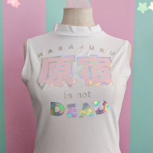 May include: White sleeveless shirt with a holographic print that says "Harajuku" and "原宿" in Japanese characters, followed by "is not DEAD".