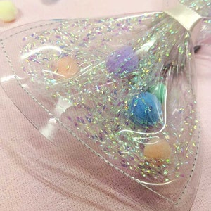 May include: A clear plastic bow with iridescent glitter and colorful pom-poms inside. The bow is on a pink surface.