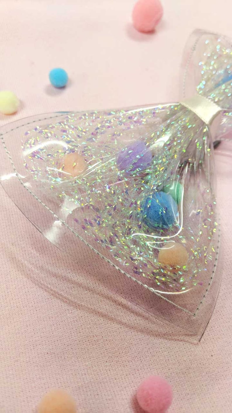May include: A clear plastic bow with iridescent glitter and colorful pom-poms inside. The bow is on a pink surface.