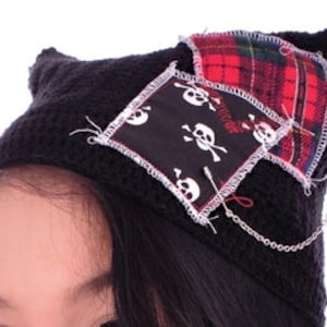May include: Black knit beanie with a cat ear detail, featuring a skull and crossbones patch and a plaid patch. A silver chain hangs from the ear.