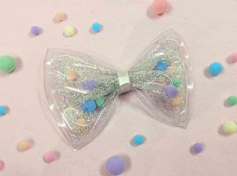 May include: A clear plastic bow with a silver glitter finish and colorful pom-poms inside.