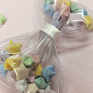 May include: A clear plastic bow with a white ribbon tied around it. The bow is filled with colorful origami stars.