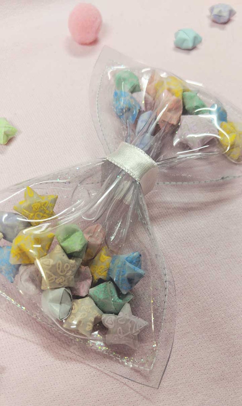 May include: A clear plastic bow with a white ribbon tied around it. The bow is filled with colorful origami stars.