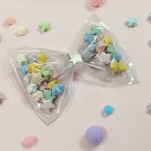 May include: A clear plastic bow with a white ribbon, filled with colorful origami stars. The bow is on a pink surface with small, colorful pom-poms scattered around it.