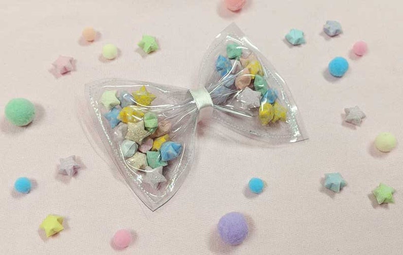 May include: A clear plastic bow with a white ribbon, filled with colorful origami stars. The bow is on a pink surface with small, colorful pom-poms scattered around it.