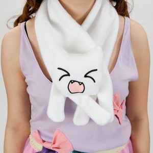 May include: White fleece scarf with a cute cat face embroidered on it. The cat has a pink mouth and black eyes. The scarf is worn by a person wearing a purple tank top.