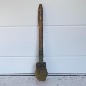 May include: A vintage paint brush with a worn wooden handle and a rounded brush head. The handle is a dark brown colour, and the bristles are a light tan. The brush shows signs of age and use.