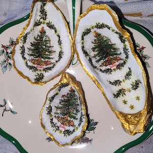 May include: Three oyster shells painted with gold trim and Christmas tree designs. Each shell features a green Christmas tree with red ornaments and a white background. The largest shell has the word "Spode" printed on the tree.