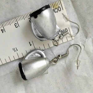 May include: A pair of silver-colored teapot earrings with black accents. The earrings have a hook closure and are shown next to a ruler for scale. The teapot design includes a handle and spout.