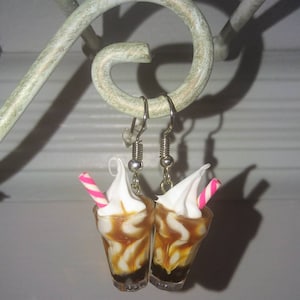 May include: A pair of dangle earrings featuring miniature milk shakes with white whipped cream, brown chocolate syrup, and pink and white striped straws.
