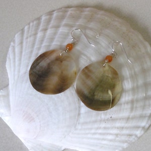 May include: A pair of handcrafted earrings featuring large, round, shell-like pendants with brown and tan hues. Each earring has a small orange bead and a silver-colored hook. The earrings are displayed on a white seashell.