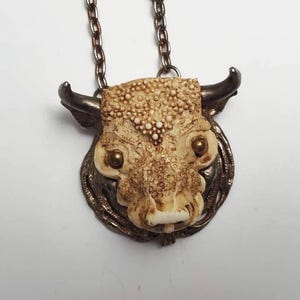May include: A silver chain necklace with a detailed, carved, bone-like bull's head pendant. The pendant has gold-colored eyes and a textured surface.