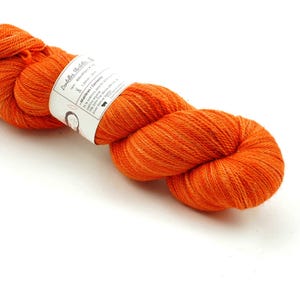 Puede incluir: Un ovillo de lana naranja con una etiqueta blanca que dice "Puddles Studio" y "Merino/Corriedale".