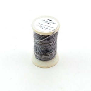 Peut inclure: Une bobine de fil gris avec une base en bois et une étiquette blanche. L'étiquette comporte du texte et des chiffres. Le fil est enroulé serré autour de la bobine, créant une forme cylindrique. Le fil semble être un mélange de gris et de marron.