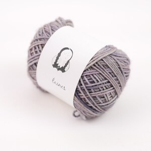 Peut inclure: Une pelote de laine violette avec une étiquette blanche sur laquelle est inscrit "laines" et une petite illustration d'une personne avec des tresses.