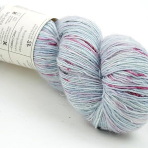 Peut inclure: Une pelote de laine bleu clair avec des mouchetures roses et violettes. La laine est bien enroulée et porte une étiquette. L'étiquette contient du texte et des symboles. La laine est probablement utilisée pour le tricot ou le crochet.
