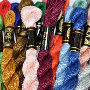 May include: A collection of DMC size 5 embroidery thread skeins in a variety of vibrant colors including yellow, green, gold, brown, pink, burgundy, orange, light blue, dark blue, peach, and silver, each wrapped with a black and gold paper label.