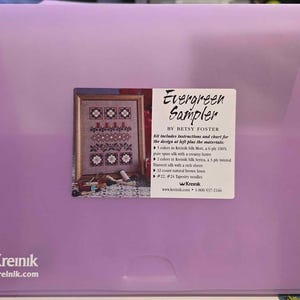 May include: A light purple plastic folder containing an Evergreen Sampler cross-stitch kit by Betsy Foster. The label displays a finished sampler design, a list of included silk threads, natural brown linen, and tapestry needles.