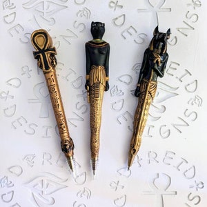 May include: Three gold-colored pens with Egyptian-themed designs. The pens feature an ankh, a cat, and a jackal.
