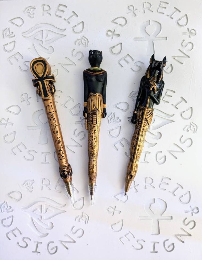 May include: Three gold-colored pens with Egyptian-themed designs. The pens feature an ankh, a cat, and a jackal.