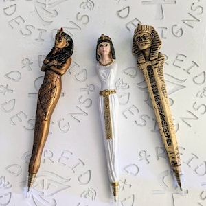 May include: Three decorative pens with Egyptian-themed figures. The pens are gold, white, and brown. The figures are of a woman with a headdress, a woman with a headdress, and a man with a headdress.