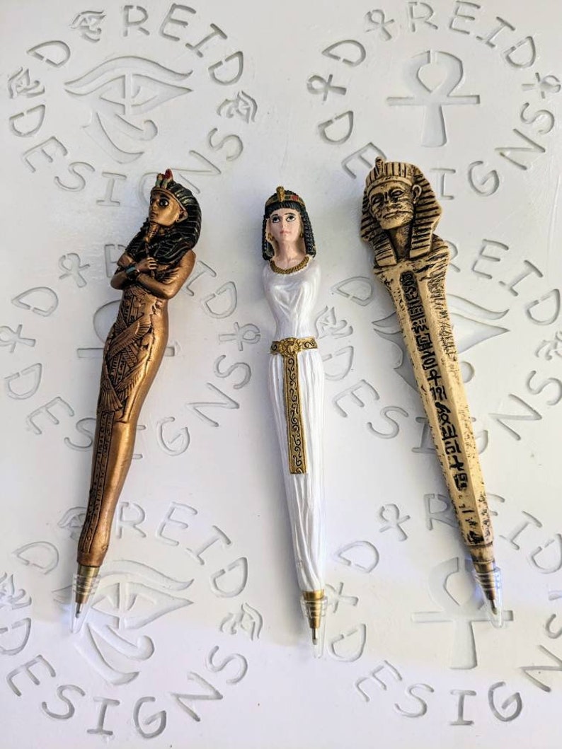 May include: Three decorative pens with Egyptian-themed figures. The pens are gold, white, and brown. The figures are of a woman with a headdress, a woman with a headdress, and a man with a headdress.