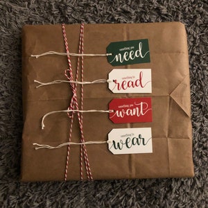 May include: A brown paper package wrapped with red and white twine and four gift tags. The tags are white with red and green lettering. The text on the tags reads "something you need", "something to read", "something you want", and "something to wear".