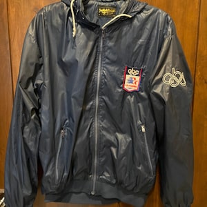 May include: Navy blue windbreaker jacket with a hood and a zipper closure. Features a patch with text and logo on the chest and a logo on the sleeve. The jacket is hanging on a wooden door.