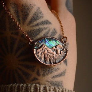May include: A copper necklace featuring a mountain scene pendant. The pendant is crafted from copper and incorporates a piece of abalone shell in the centre. The abalone shell displays iridescent hues of green, blue, and purple.