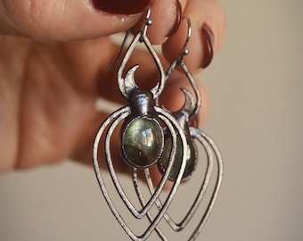 May include: Silver-toned earrings with a layered heart design. Each earring features a central, oval-shaped, iridescent stone. The earrings are held up by a silver hook.