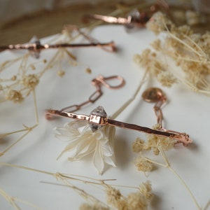 May include: A delicate rose gold bracelet with a hammered finish and a clear crystal charm. The bracelet is displayed on a white surface with dried flowers.