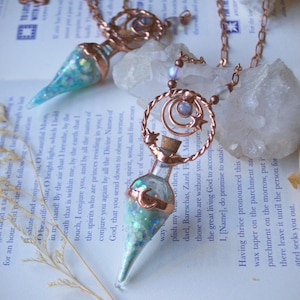 May include: Two copper-colored necklaces with glass vial pendants filled with blue and green glitter. Each pendant is topped with a copper crescent moon and star design. The necklaces rest on a book with text.
