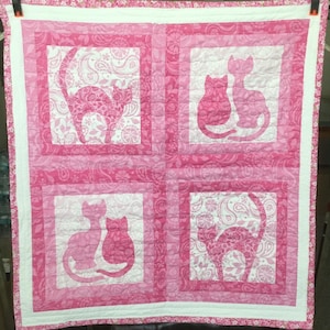 May include: A pink and white quilt featuring four squares with cat silhouettes. The cats are in various poses and filled with a paisley pattern. The quilt has a white border and is made of fabric.