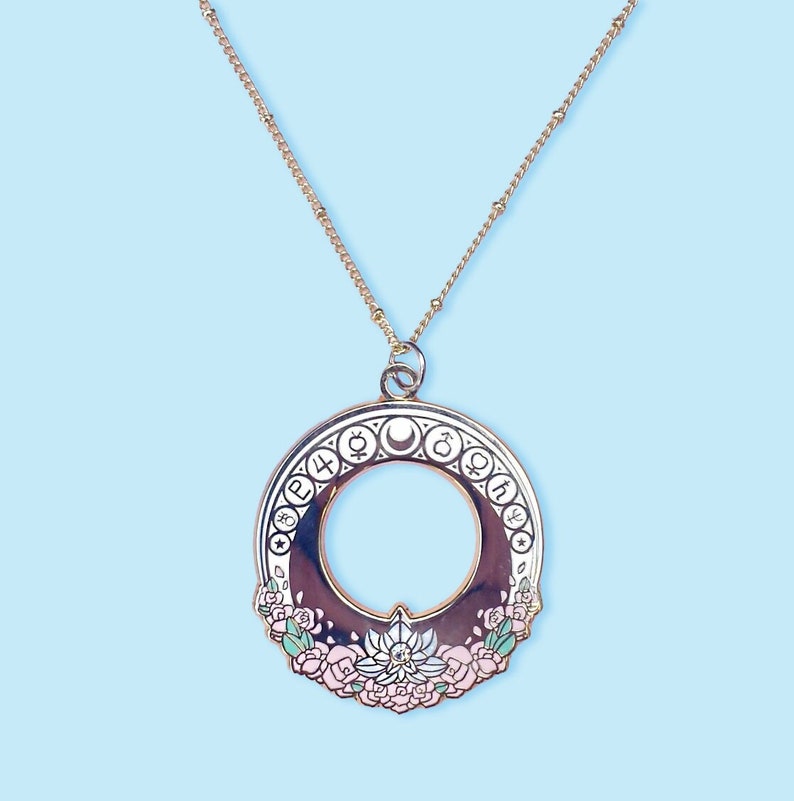 A gold tone necklace chain with an art nouveau style sailor moon gold and enamel pendant featuring the planetary symbols on the outside. The bottom has flowers and a silver crystal lotus with a gem. The middle has a cutout with a gold crescent moon.