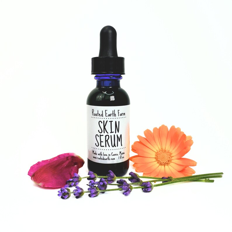 May include: A small, dark glass bottle with a dropper top, labeled "Rooted Earth Farm Skin Serum". The bottle is surrounded by lavender flowers and a single orange flower.
