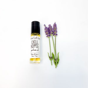 Puede incluir: Una peque&ntilde;a botella de vidrio con un aplicador de rodillo lleno de l&iacute;quido amarillo. La botella tiene una etiqueta blanca con el texto "Wilder Earth Farm CHILL HIPPIE perfume oil Made with love in Casco, Maine". Un tallo de lavanda con flores moradas est&aacute; al lado de la botella.
