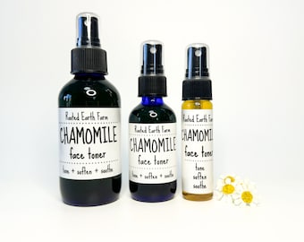 May include: Three glass bottles with spray tops containing chamomile face toner. The bottles are labeled "Rooted Earth Farm Chamomile Face Toner" and list the benefits as "tone + soften + soothe". The bottles are arranged in order of size from left to right.  A few white daisy flowers are in the bottom right corner of the image.