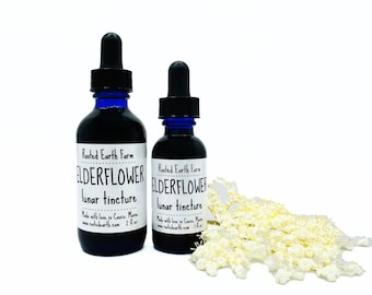 May include: Two dark blue glass bottles with black droppers, labelled "ELDERFLOWER lunar tincture," are displayed. A cluster of small, cream-coloured flowers is to the right. The bottles are 59 ml and 30 ml.